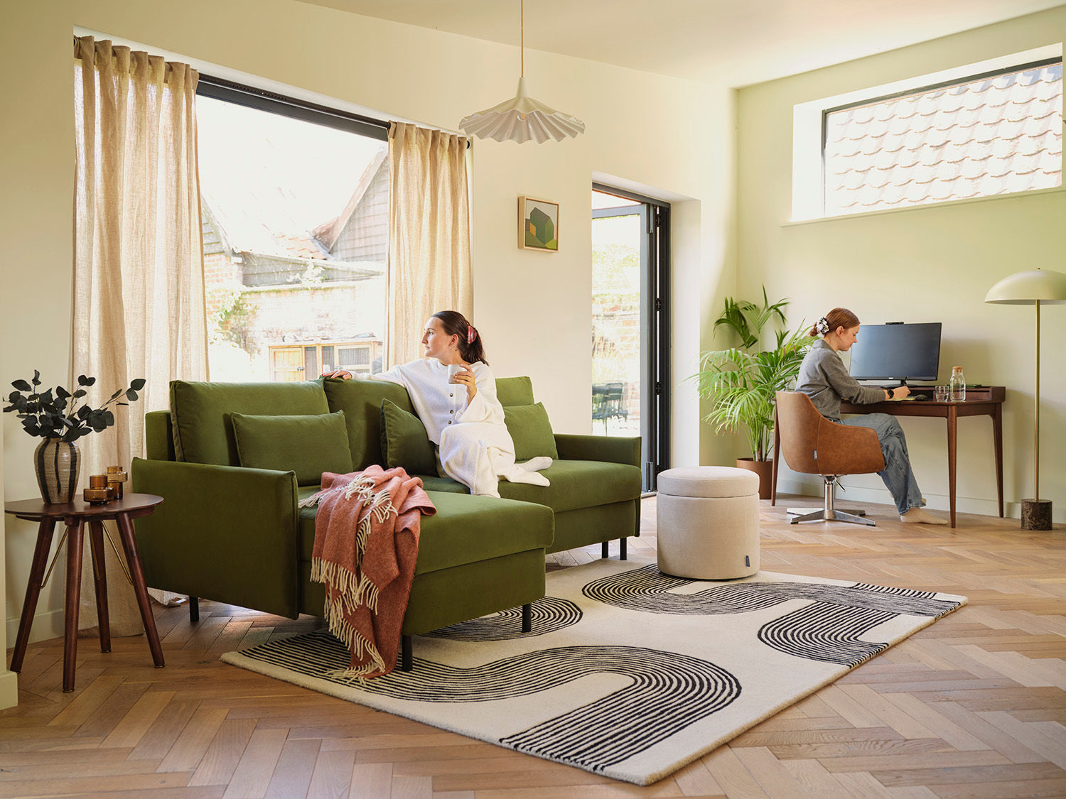 Living room with green sofa, rug, and a woman working at a desk.